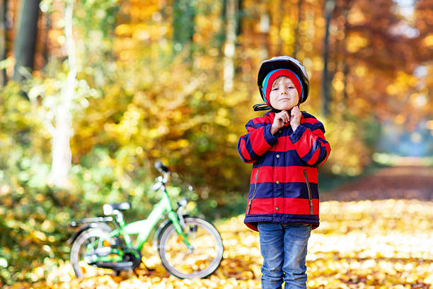 Little kid boy in colorful warm clothes in autumn forest park with a bicycle. Active child putting safe helmet before cycling on sunny fall day in nature. Safety, sports, leisure with kids concept.