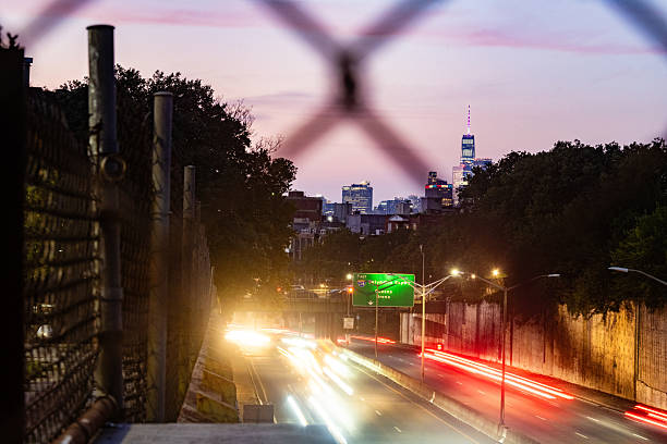 This is a long exposure photograph of traffic on a highway in Brooklyn, and the New York City skyline with the Freedom Tower illuminated at sunset, and seen through a chain-link fence.