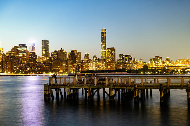 This is a photograph of Long Island City docks in Queens with the Manhattan skyline in the background. The buildings are illuminated above the East River on a summer night in New York City.