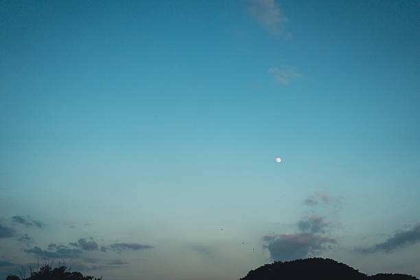Small disk of the moon against a blue sky and a mountain in twilight.