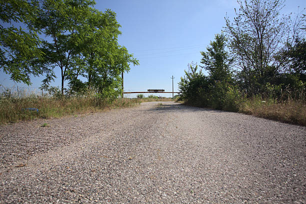 Worn paved road blocked by a rusty bar with the main passage framed by trees on a sunny day