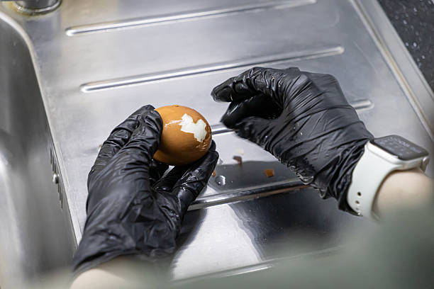 istock Hands in black gloves meticulously peeling a brown hard-boiled egg over a clean stainless steel kitchen sink, ensuring safe food handling and preparation hygiene 2237889014