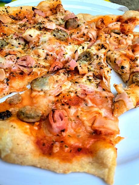 Overhead view of a freshly baked homemade pizza with thin crust, topped with ham, mushrooms, tomato sauce, melted cheese, and Italian herbs. Made with a 65% hydration dough and cold-fermented for 40 hours. Authentic and rustic food presentation on a white plate.