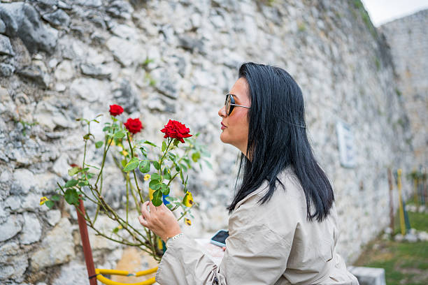 Close-up of woman with long black hair and sunglasses looking at blooming red roses next to an old stone wall. She wears a beige trench coat and holds a smartphone in her hand while bending slightly towards the flowers.