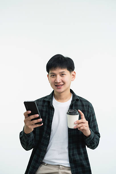istock Young man holding smartphone and coffee cup, smiling, standing against white background, enjoying modern lifestyle, casual style, trendsetting expression 2237968526