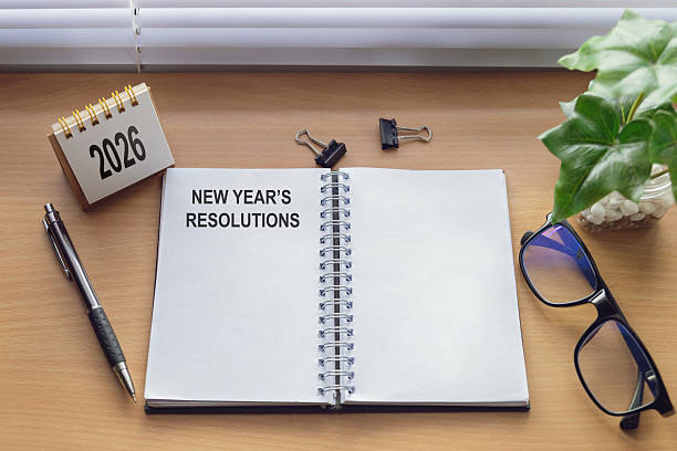 istock Flat lay of open notebook title NEW YEAR'S RESOLUTIONS on wooden desk with copy space, 2026 calendar, pen, glasses, binder clips, and green plant; goal setting, New Year planning, productivity concept 2237972236