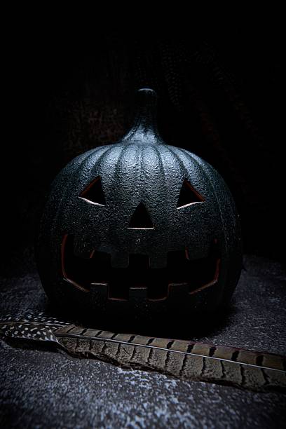 Dark Halloween still life with a carved jack-o'-lantern in moody low-key light and a pheasant feather on a textured surface, spooky season background.