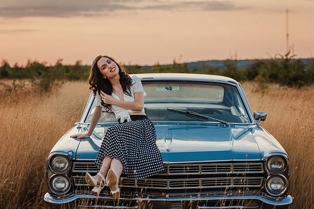 Pretty young woman with retro clothes sitting on a classic coupe's hood stock photo