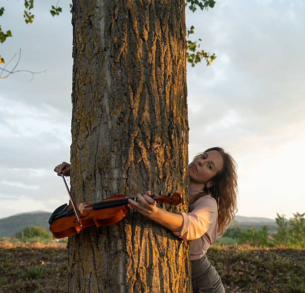 A woman holding a tree and playing with a violin outdoor.