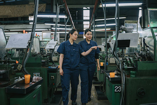 Two Asian female workers in blue uniforms inspecting and discussing machine operations on a factory floor, representing teamwork, precision, and gender diversity in technical industries.