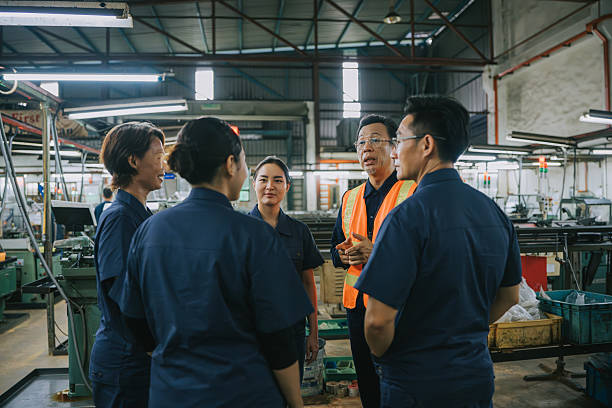 Malay Supervisor in a safety vest conducting a team briefing with factory operators inside an industrial plant, representing leadership, teamwork, and workplace communication.