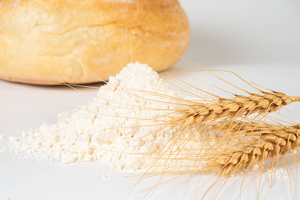 Pile of flour beside dried wheat stalks with a round cheese in the backdrop, highlighting essential cooking ingredients.