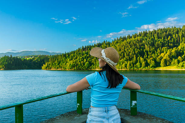 Woman in a straw hat and blue t-shirt leans on a railing, enjoying a wide view of the calm lake and green forested hills. The scene radiates relaxation, harmony with nature, and summer leisure.