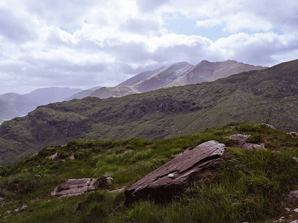 Expansive Irish landscape. Lush green hills under a soft sky. Rocks in foreground add texture.