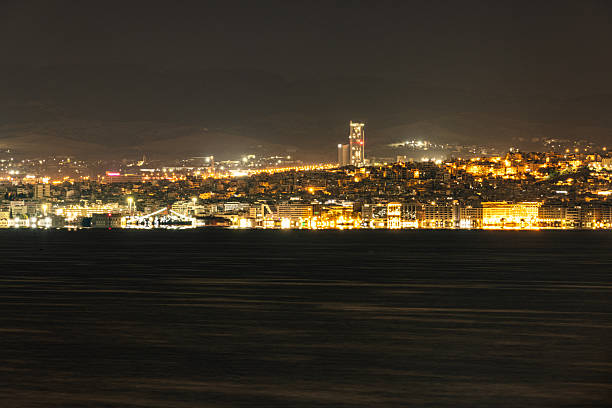 Night view of Thessaloniki waterfront and commercial port in Northern Greece. The illuminated cityscape includes seafront buildings, modern high-rise structures, and the glowing skyline along the Thermaic Gulf.