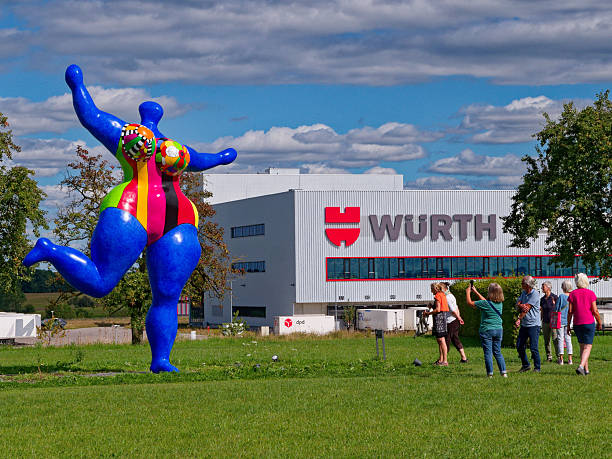 Künzelsau, Germany - August 31, 2025: Public sculpture garden of the Museum Würth 2 in Künzelsau on a summer's day. A group of visitors view and photograph a colorful Nana sculpture by artist Niki de Saint Phalle. In the background stands a building belonging to the Würth company.
