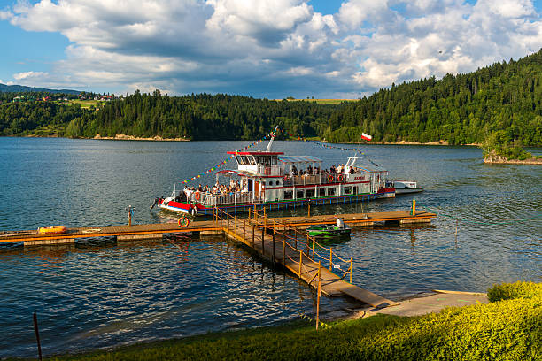 Czorsztyn Lake, Poland – August 5, 2025: A festive steamboat filled with tourists arrives at the pier. The vessel, adorned with colorful flags and the Polish flag, contrasts with the green hills and calm blue water surrounding it.