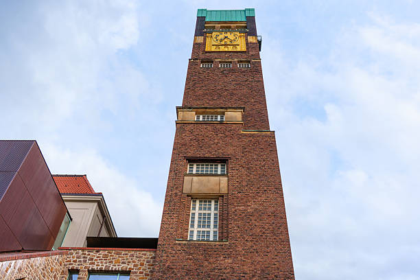 Darmstadt, Germany - June 5, 2025: Close-up view of the Wedding Tower in Darmstadt, Germany, showcasing its brick structure and prominent clock, framed by contemporary buildings under a cloudy sky.