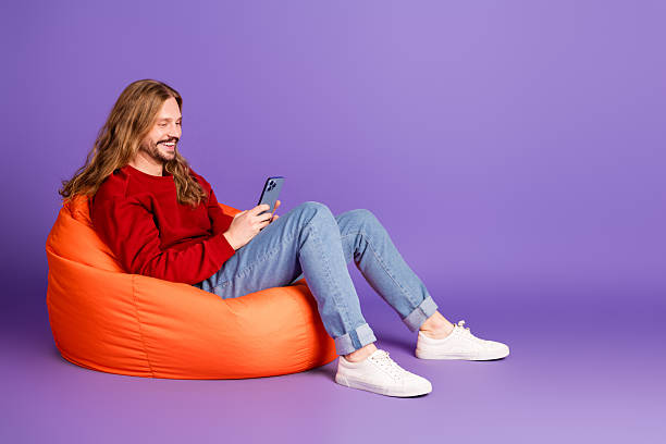 istock Young handsome man relaxing on an orange bean bag while using his smartphone against a purple background 2238013910