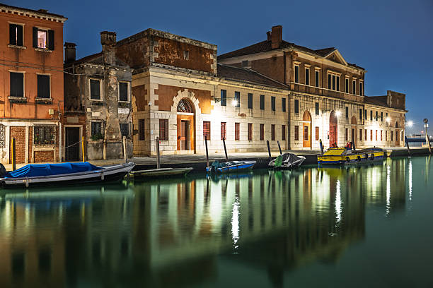 Long exposure captures the smooth water of the lagoon and boats drifting by illuminated windows of Campus San Giobbe in Venice at night.