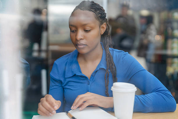 A young African woman with braided hair, wearing a blue shirt, sits at a table. She's focused and writing in a notebook with a pen. A disposable coffee cup sits next to her.
