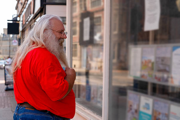 An older man with a long white beard and glasses stands on a sidewalk in England. He is wearing a red shirt and jeans and looking into the window of a shop.