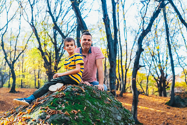 A father and his young son sit together on a mossy rock in a sunlit autumn forest. Surrounded by colorful leaves and tall trees, they enjoy a peaceful family moment in nature, reflecting bonding and outdoor adventure.