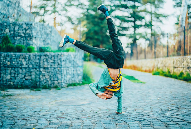 A cheerful young boy performs a one-handed handstand on a cobblestone path in a scenic park, showing balance, energy, and agility. His striped shirt and enthusiastic pose capture childhood fun, movement, and the joy of playful outdoor activities.