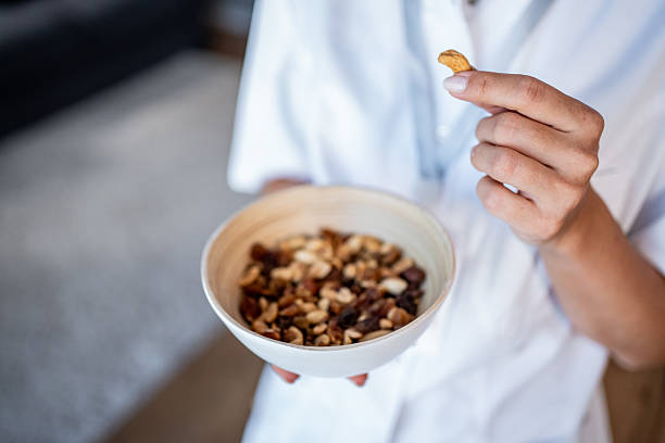 Adult woman wearing a white coat holding a bowl of trail mix and picking a cashew, showcasing healthy eating, balanced diet, and nutritious snacking at work