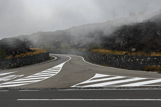 Curved mountain road with fresh markings and stone walls on the slopes of Mount Etna in Sicily, Italy. Mist and volcanic rocks create a dramatic atmosphere.