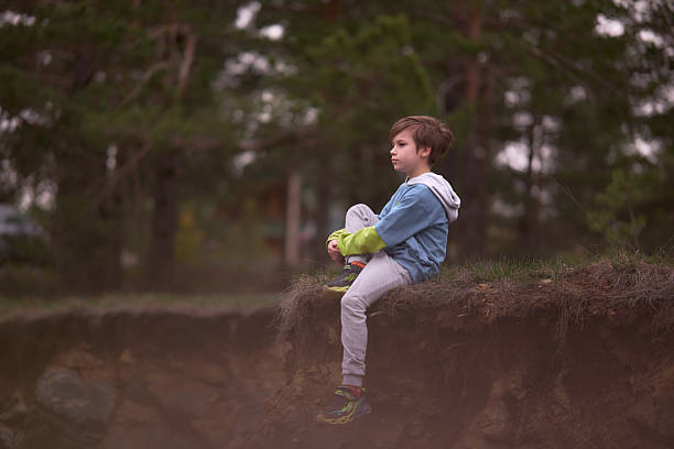 Young boy sitting outdoors on a dirt ledge, daydreaming and looking into the distance.