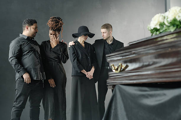 Group of young adult and middle aged multiethnic men and women standing together mourning near closed casket during funeral ceremony, showing support and comforting each other