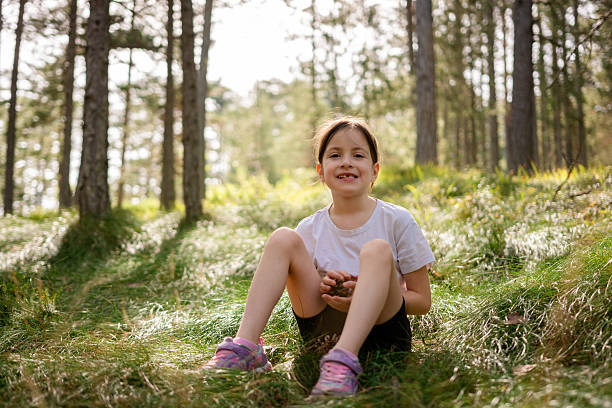 Young girl smiling at the camera, holding small pinecones and sitting on green grass in a forest on a sunny day