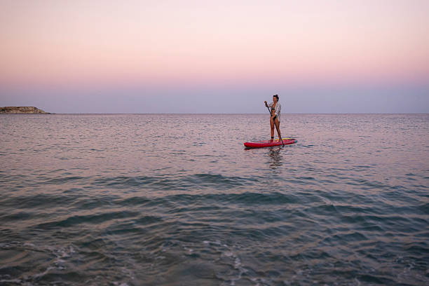 A woman paddleboards on calm ocean waters during a peaceful sunset. The soft pastel sky and gentle waves create a serene scene.