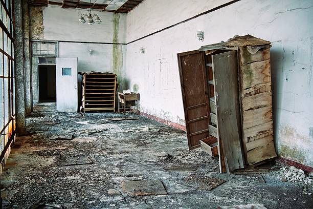 This evocative image portrays the haunting stillness of a forgotten office interior, where cracked white walls and a sagging ceiling chandelier frame an open, weathered wooden wardrobe spilling drawers amid dusty rubble and toppled shelves. Soft light filters through tall, rusted metal windows onto the debris-littered concrete floor, casting long shadows that whisper of time's relentless passage and urban neglect. The moody composition captures themes of decay, nostalgia, and quiet desolation, ideal for editorial storytelling, atmospheric advertising, post-apocalyptic concepts, or conceptual photography in films, books, or lifestyle campaigns.