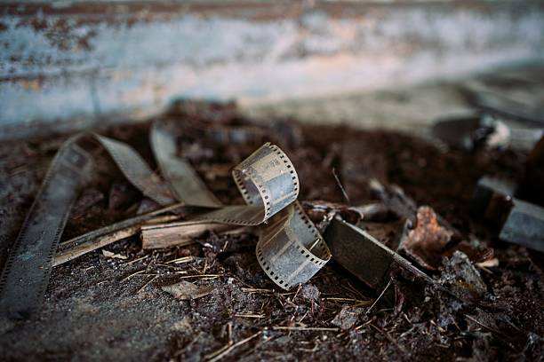 This textured close-up captures weathered strips of old 35mm film unspooled and tangled among scattered wood shavings, rusted metal scraps, and dusty concrete in a forgotten industrial space, with faint sunlight highlighting the perforated edges against a corroded wall backdrop. The nostalgic patina evokes cinema history, creative decay, and timeless storytelling, ideal for film-themed visuals, retro advertising, urban exploration concepts, or atmospheric editorial in movies, books, or lifestyle campaigns.