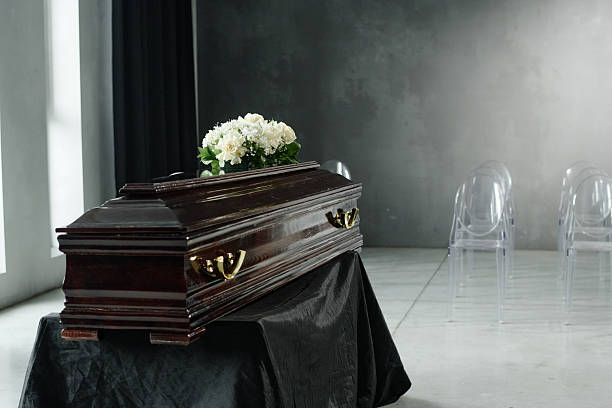 Closed wooden coffin with flower arrangement resting on top, positioned on black cloth in modern funeral setting with empty transparent chairs in background