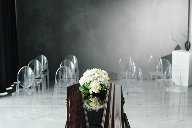 Closed wooden coffin with floral arrangement standing in front of empty transparent chairs in modern funeral setting, reflecting somber memorial ceremony arrangement