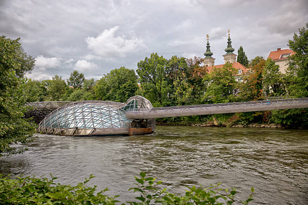 Graz, Austria – July 22, 2025: The Murinsel (Island in the Mur), a floating steel platform and pedestrian bridge designed by Vito Acconci in 2003, spans the Mur River with Schlossberg hill and surrounding architecture in the background.