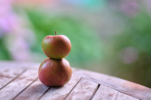 A close-up image featuring three small apples in shades of red and green sitting on a weathered wooden surface, with a lush and blurred natural background adding to the serene outdoor ambiance.