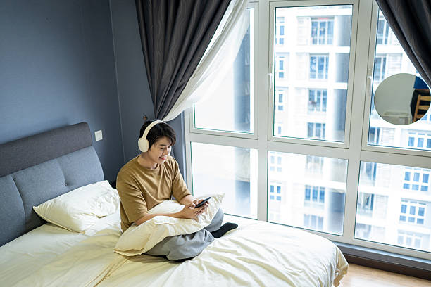 istock Young Asian man sitting on a bed with headphones on, using a language-learning app on his phone. 2238086693