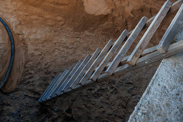 A weathered wooden ladder descends into a manually worked pit with sandy, uneven walls. A black hose is partially visible on the left side.