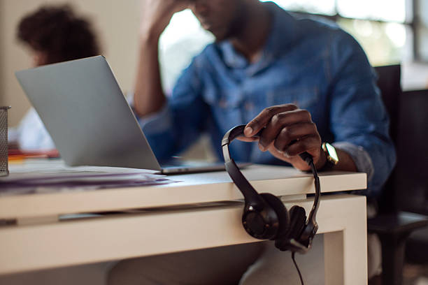 A focused individual sits at a desk using a laptop computer, holding headphones in one hand, indicating workspace activity