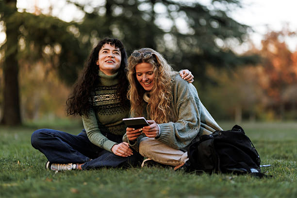 Friends enjoying a warm afternoon together in a park while sharing a tablet and laughing amidst nature's vibrant colors