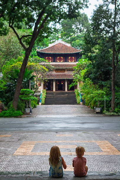 istock Two children sit on steps facing a traditional Vietnamese temple surrounded by lush tropical greenery in Saigon. Peaceful moment in cultural setting, perfect for family travel, and lifestyle themes. 2238109174