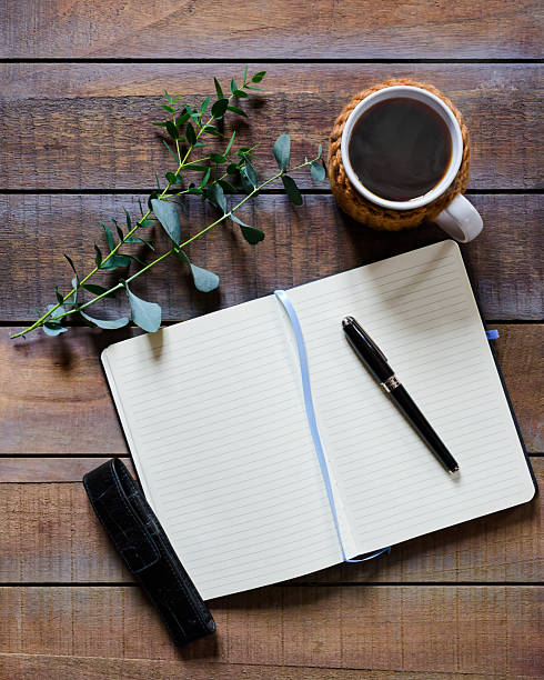 A journal lies open on a rustic wooden table, with a pen resting on its pages. A cup of black coffee and a sprig of greenery add to the calm, reflective scene. Keeping a diary can add to a feeling of calmness and relaxation. Directly above, Copy space for text - Concepts and ideas, healthy lifestyle
