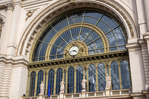 Architectural detail of Keleti Station showing decorative clock, statues, and arched glass window reflecting sky.