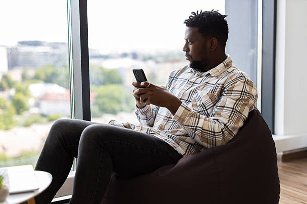 istock Man Sitting on a Beanbag Using Smartphone and Laptop by a Bright Window 2238114383
