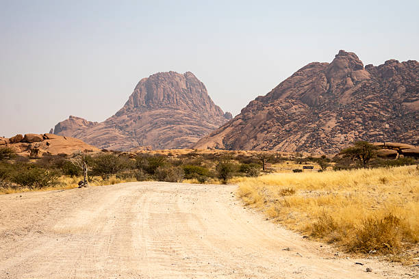 Granite peaks and rugged desert landscape at Spitzkoppe Community Campsite, Namibia. Golden grassland with scattered acacia trees contrasts against dramatic rocky mountains under a clear sky.