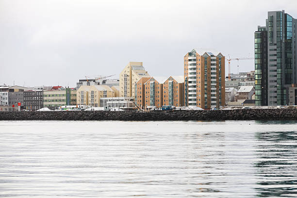 Reykjavik, Iceland, a tranquil harbor scene along a rocky breakwater, featuring colorful, modern apartments and urban buildings reflected in calm water under a pale, overcast sky